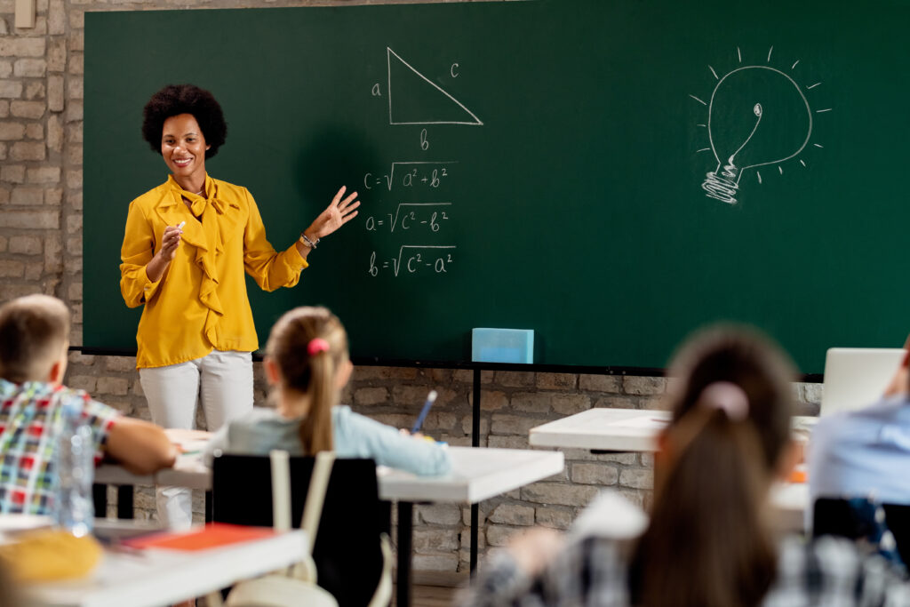 Happy African American math teacher explaining lecture on blackboard in the classroom.
